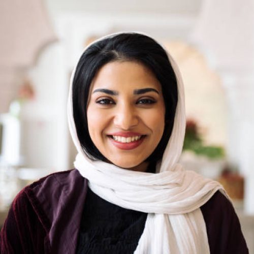 Head and shoulders portrait of woman with black hair wearing abaya, white headscarf, and smiling at camera in hotel lobby with Islamic architectural design.